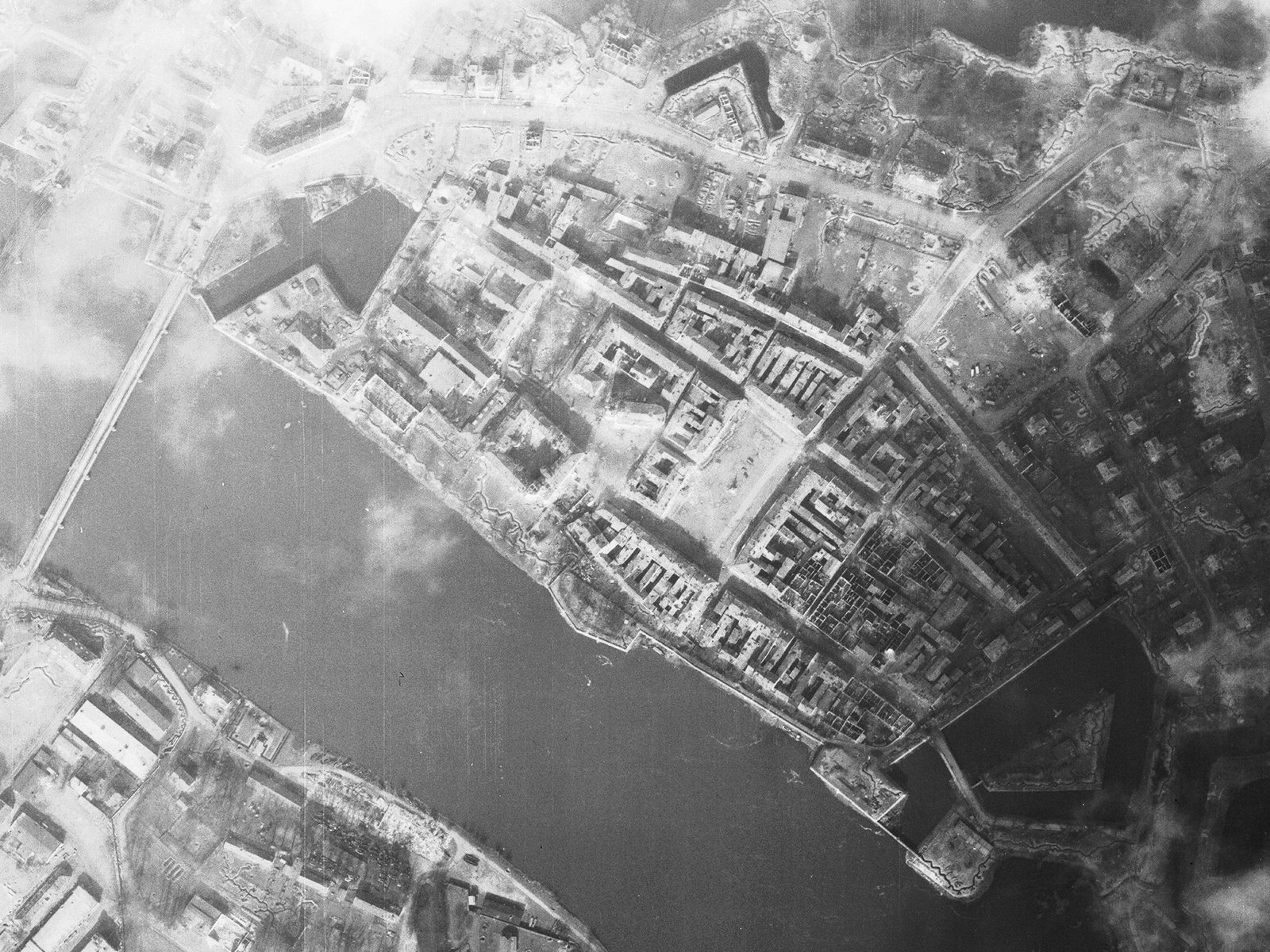 Black-and-white aerial photograph of the Altstadt of Küstrin on 18 March 1945: roofless blocks around the open market square, trenches along the Oder bank, the bastions at both ends of the town, patches of cloud.