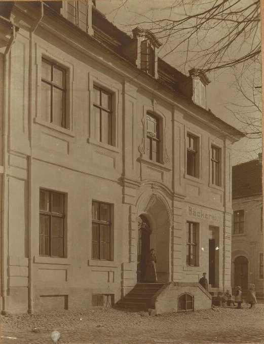 Two-storey house with a carved doorway and a sign reading Bäckerei; a woman stands in the door and children stand at the corner.