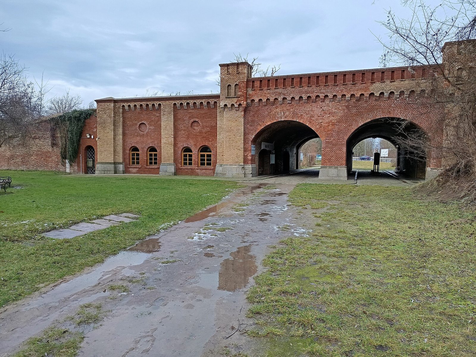 The restored Berliner Tor: a brick gatehouse with two arches and small towers, a wet cobbled road leading up to it, January 2025.