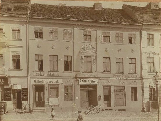 Three-storey plastered house with shop signs reading Handschuhfabrik and Zahn-Atelier; a girl and a boy stand in the street.