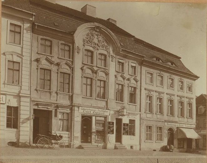 Row of baroque houses with an ornate curved gable; a carriage stands by a shop sign for colonial goods, and a bakery with an awning is at the far end.