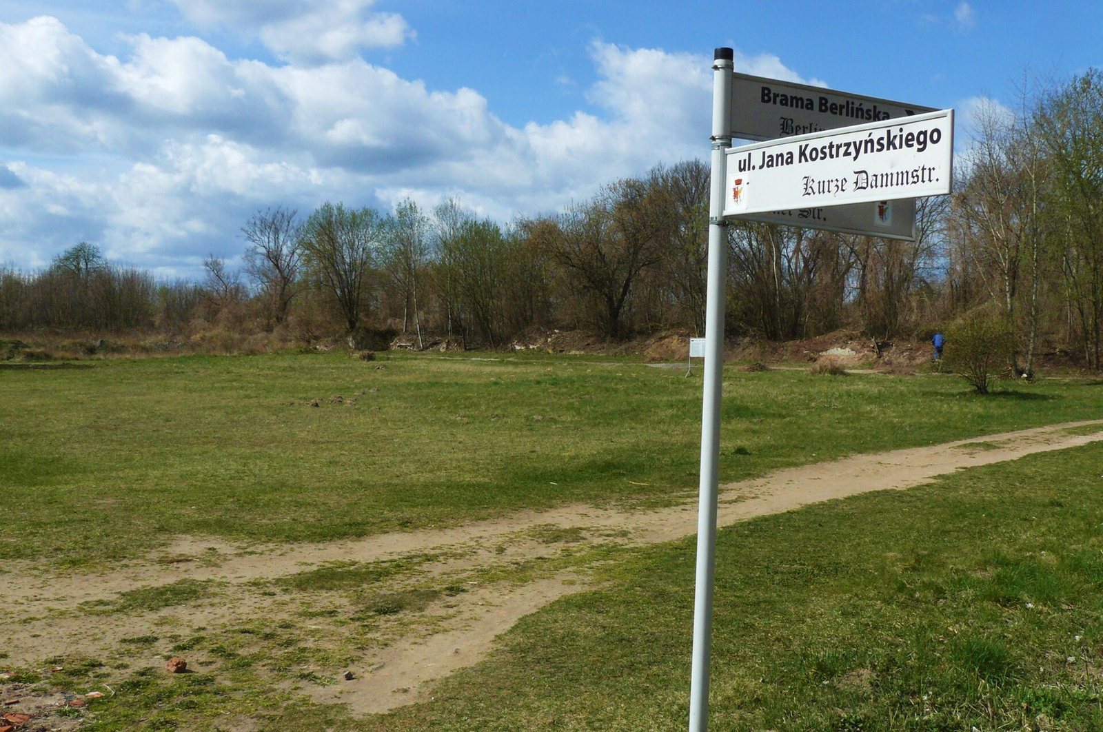 A street sign in an empty field: ul. Jana Kostrzyńskiego, with Kurze Dammstr. below it in old German lettering. A worn path crosses the grass.
