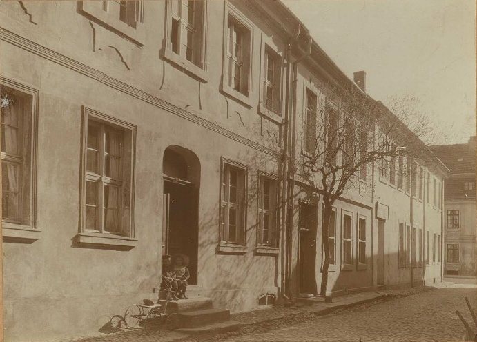 Quiet cobbled street of plastered houses; two small children sit on a doorstep beside a child's tricycle.