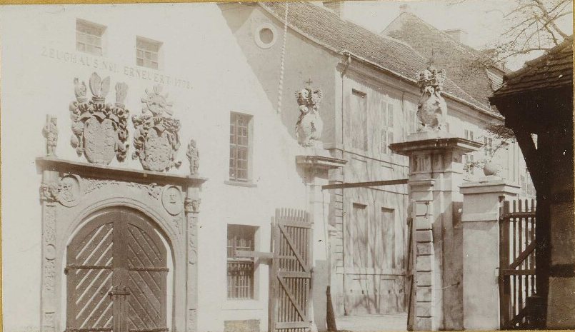 White arsenal building with a large portal under two carved coats of arms and a wooden gate with statues on its piers.